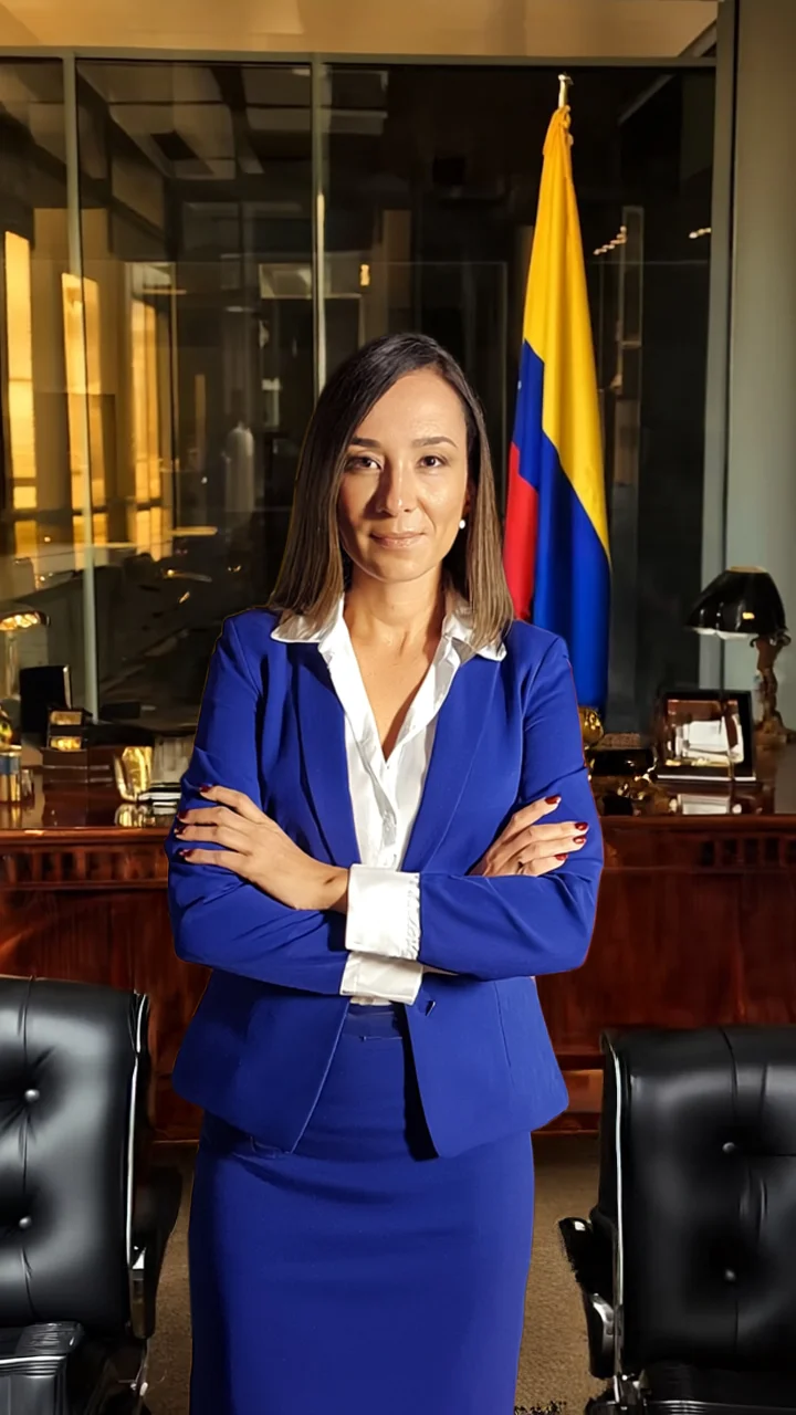 Professional woman in a blue suit stands with arms crossed in an office, a Colombian flag in the background.