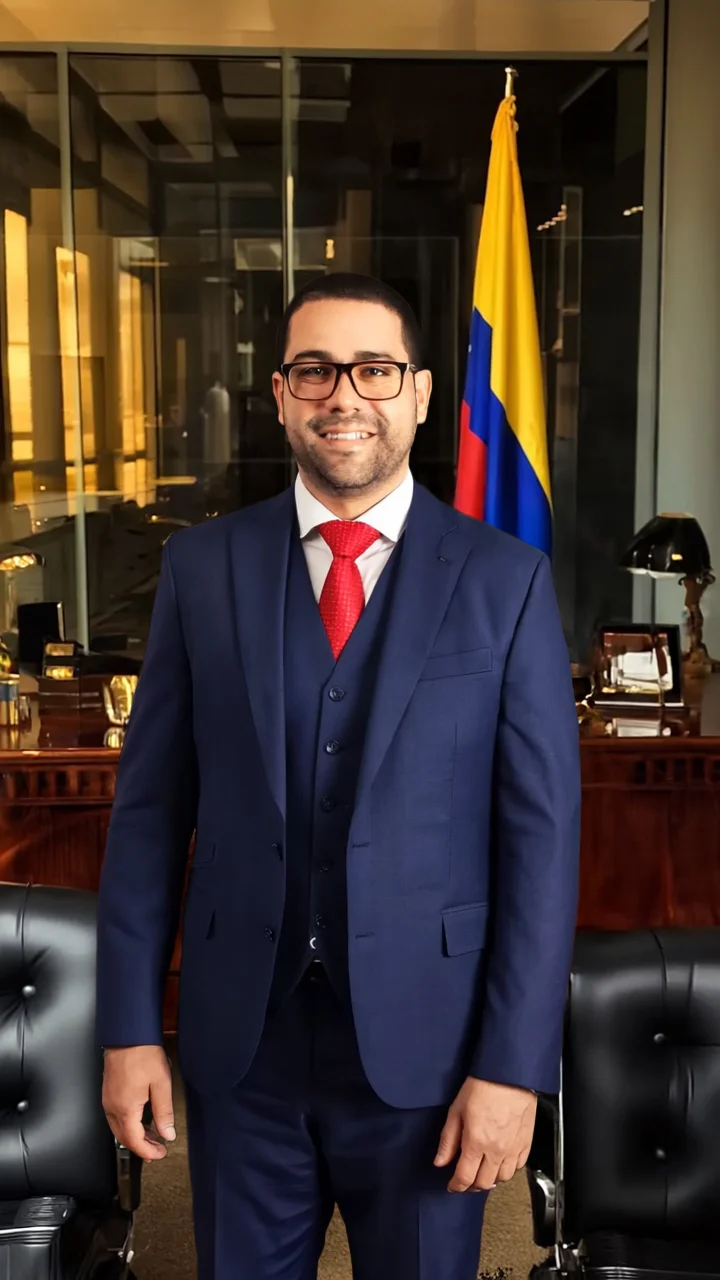 Man in a navy suit and red tie standing in an office with a Colombian flag in the background.