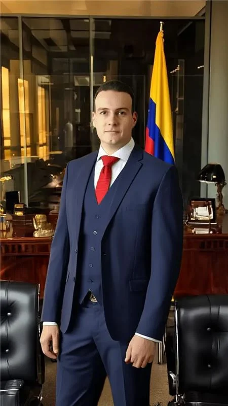 Man in a navy suit and red tie standing in an office with a yellow-blue-red flag in the background.
